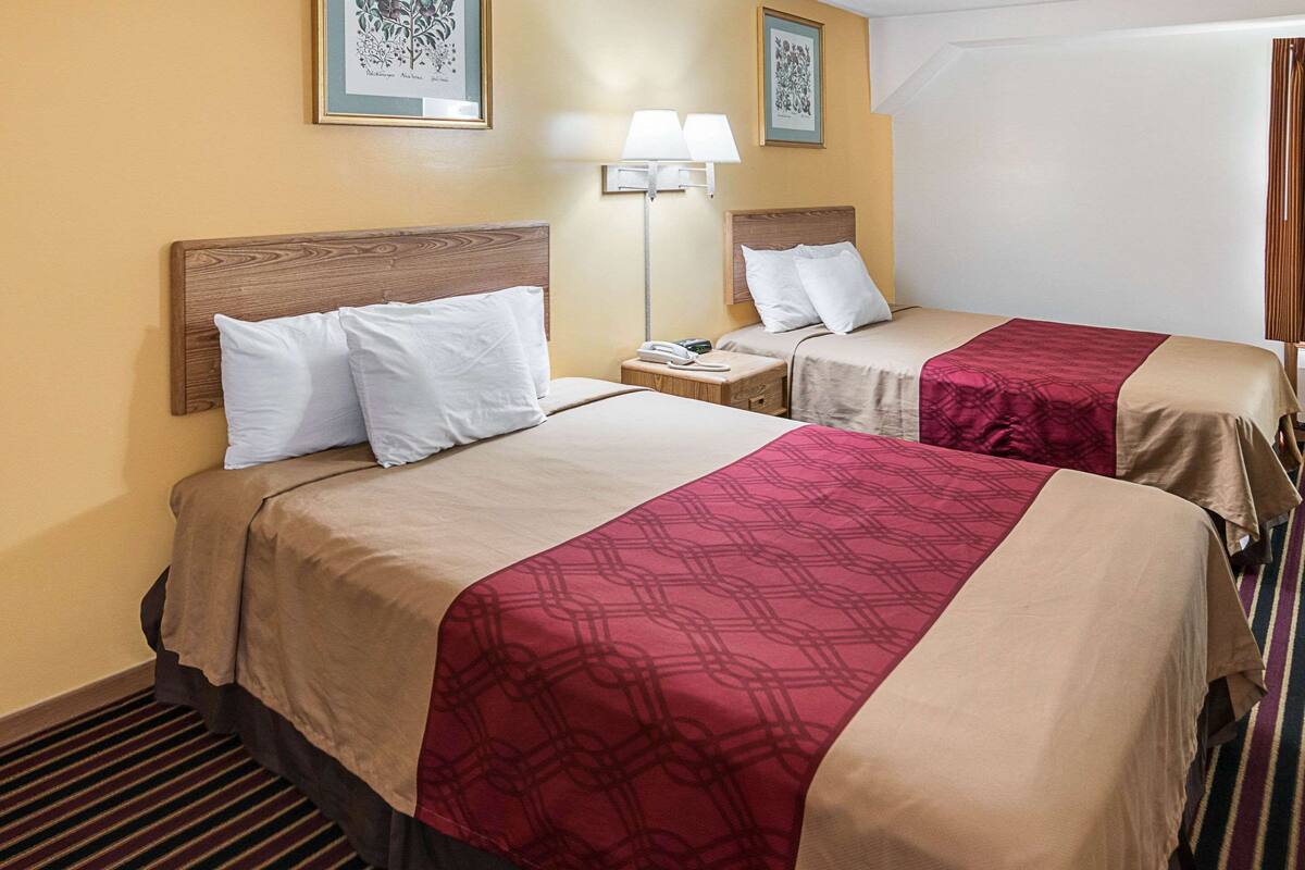 A hotel room with two beds, each with a wooden headboard, beige bedding, and a red patterned bedspread. There is a nightstand with a lamp between the beds, and the walls are painted yellow.