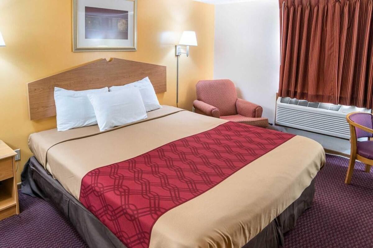 A hotel room with a bed, a chair, a lamp, a window with curtains, and an air conditioning unit.