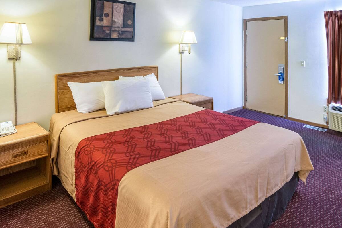A hotel room with a bed, two bedside tables, a nightstand, a wall-mounted telephone, two wall lamps, and a door.