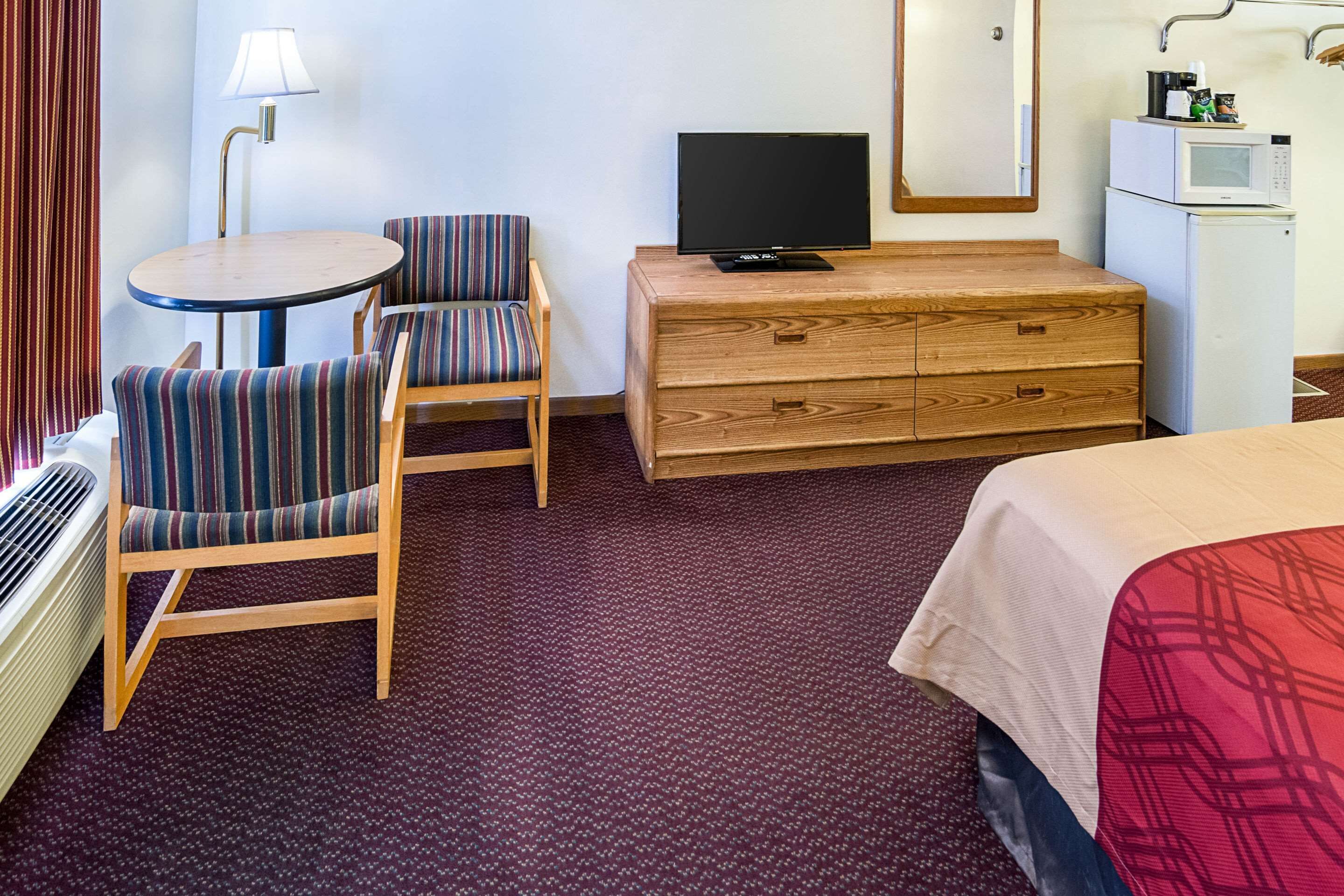 A hotel room with a bed, a TV on a wooden dresser, a striped chair, a small round table, and a microwave on a cabinet.