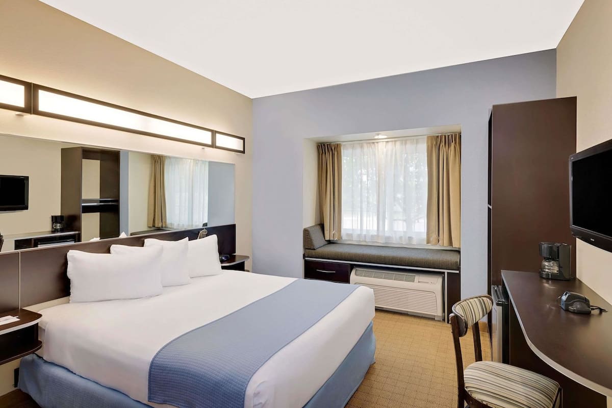 A hotel room with a large bed, a desk with a chair, a television, a window with curtains, and a built-in heater.