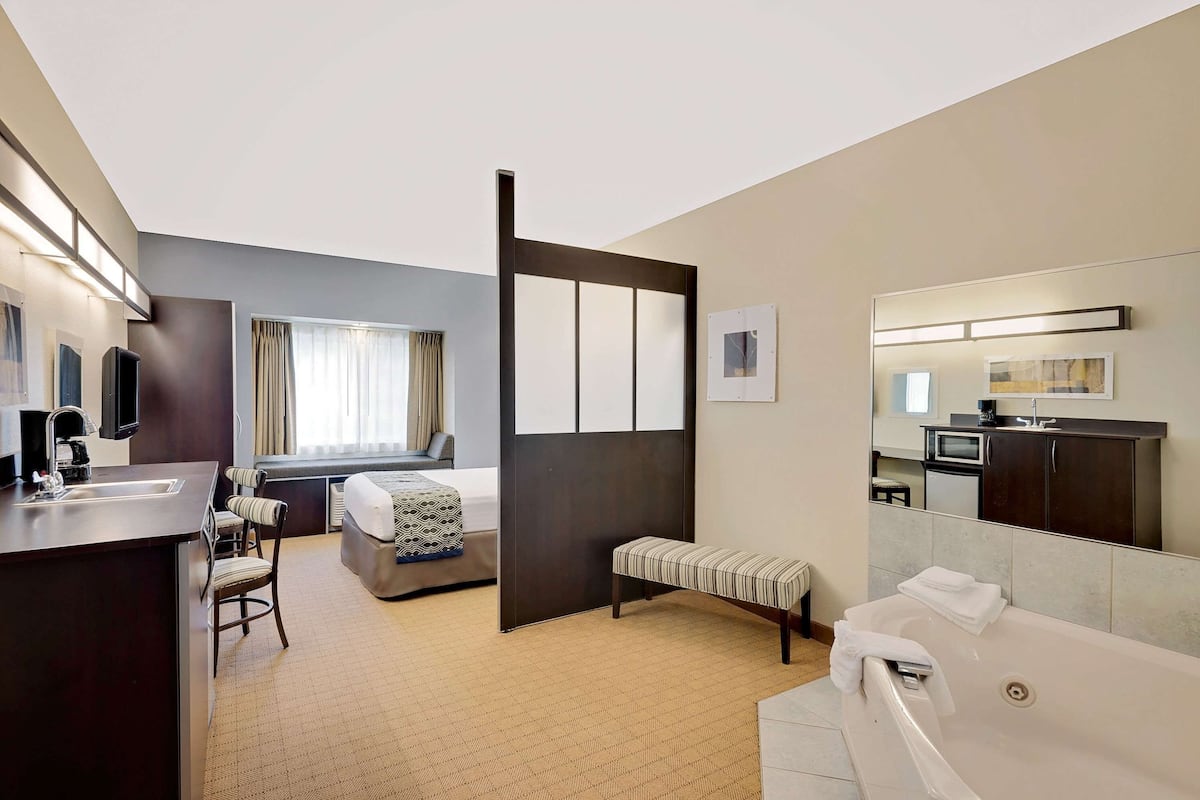 A hotel room with a bed, a desk with a chair, a bench, a bathtub, a mirror, and a TV.