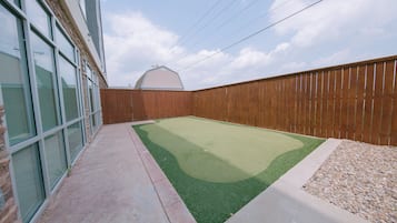 A backyard with a green artificial turf, a wooden fence, and a building with large windows.