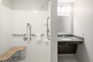 A bathroom with white tiles, a shower with grab bars, a bench, and a sink with a granite countertop.