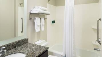 A bathroom with a granite countertop sink, a mirror, a towel rack with folded towels, a shower area with a white curtain, and a bathtub.