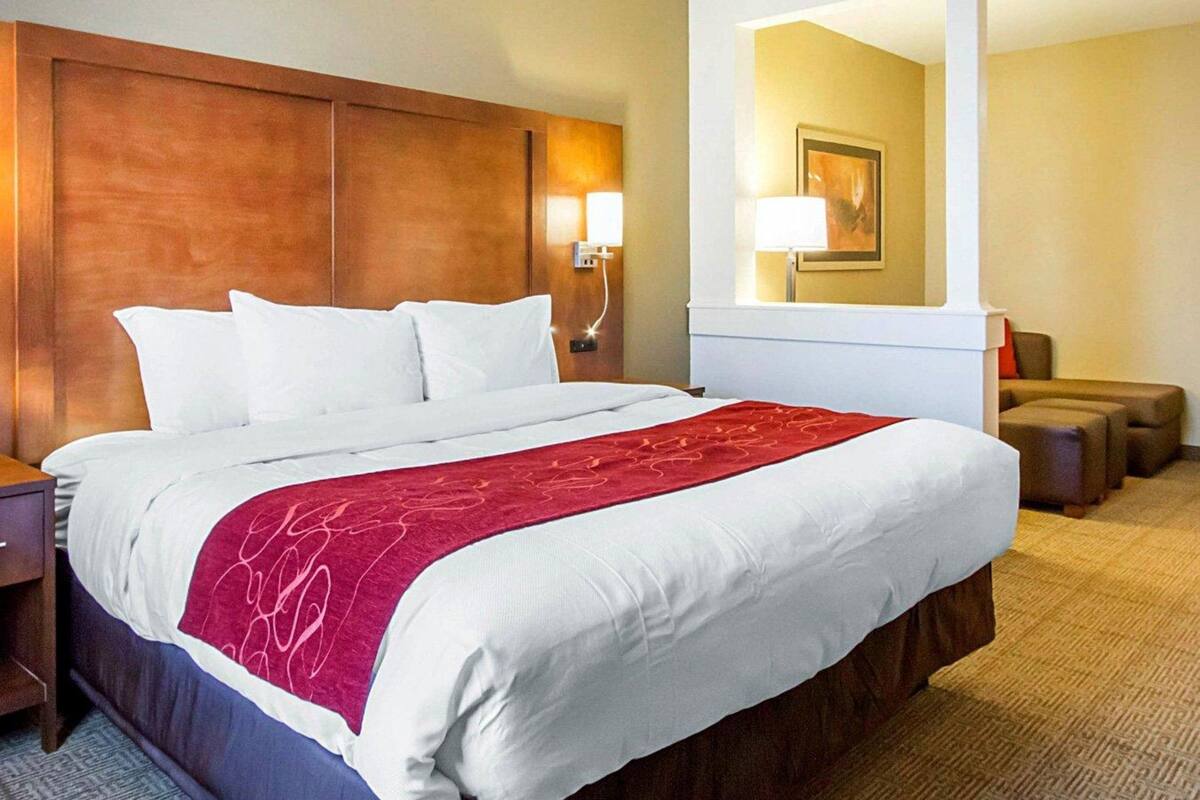 A hotel room with a large bed, a wooden headboard, a nightstand, a lamp, a mirror, and a seating area with ottomans.
