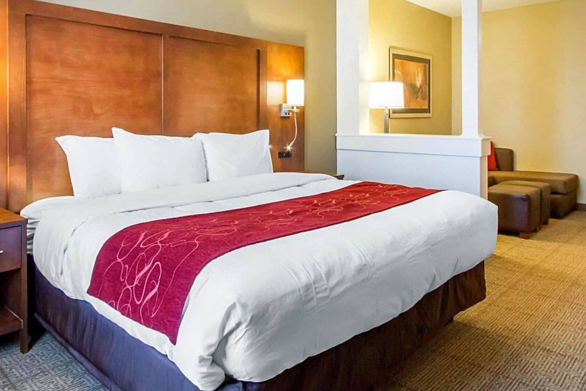 A hotel room with a large bed, a wooden headboard, a nightstand, a lamp, a mirror, and a seating area with ottomans.