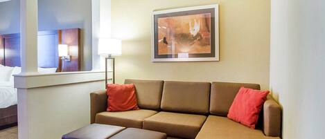 Habitación de hotel con sofá marrón, dos almohadas rojas, una lámpara de pie y un cuadro abstracto enmarcado en la pared.