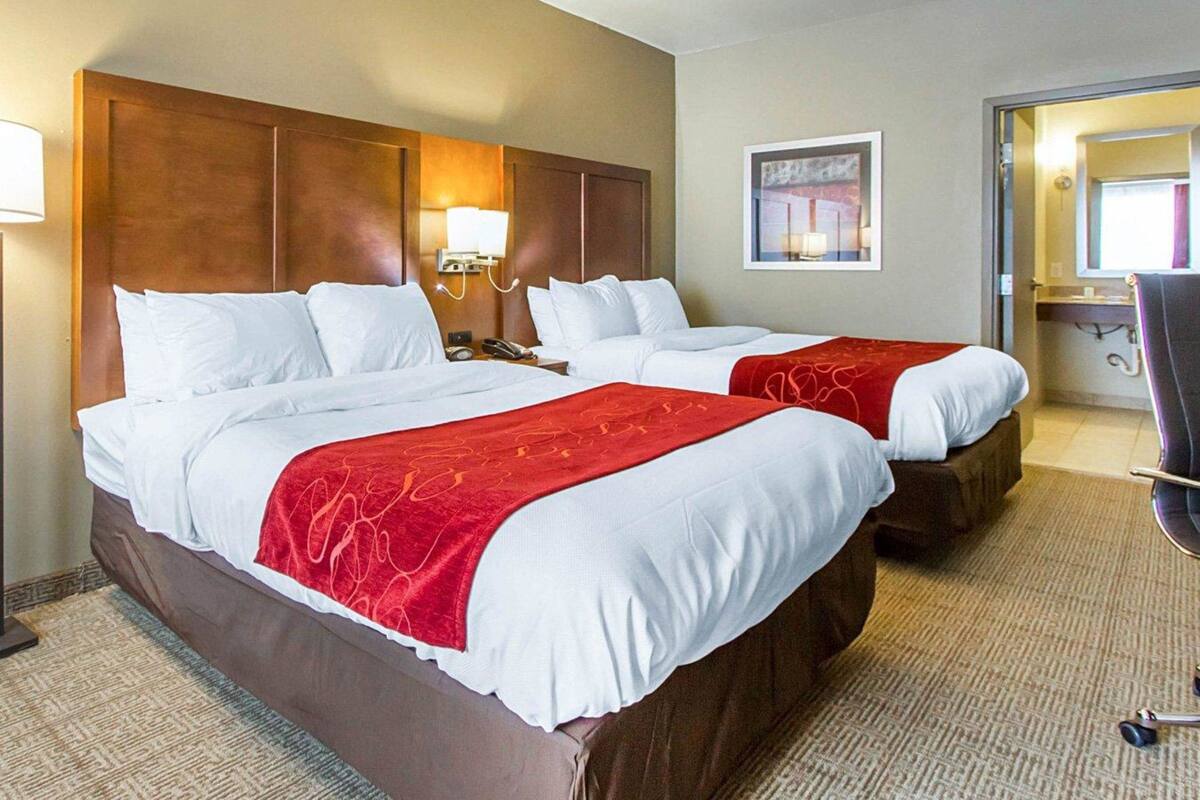 A hotel room with two beds, a wooden headboard, a nightstand with a lamp, a framed picture on the wall, and a bathroom visible through an open door.