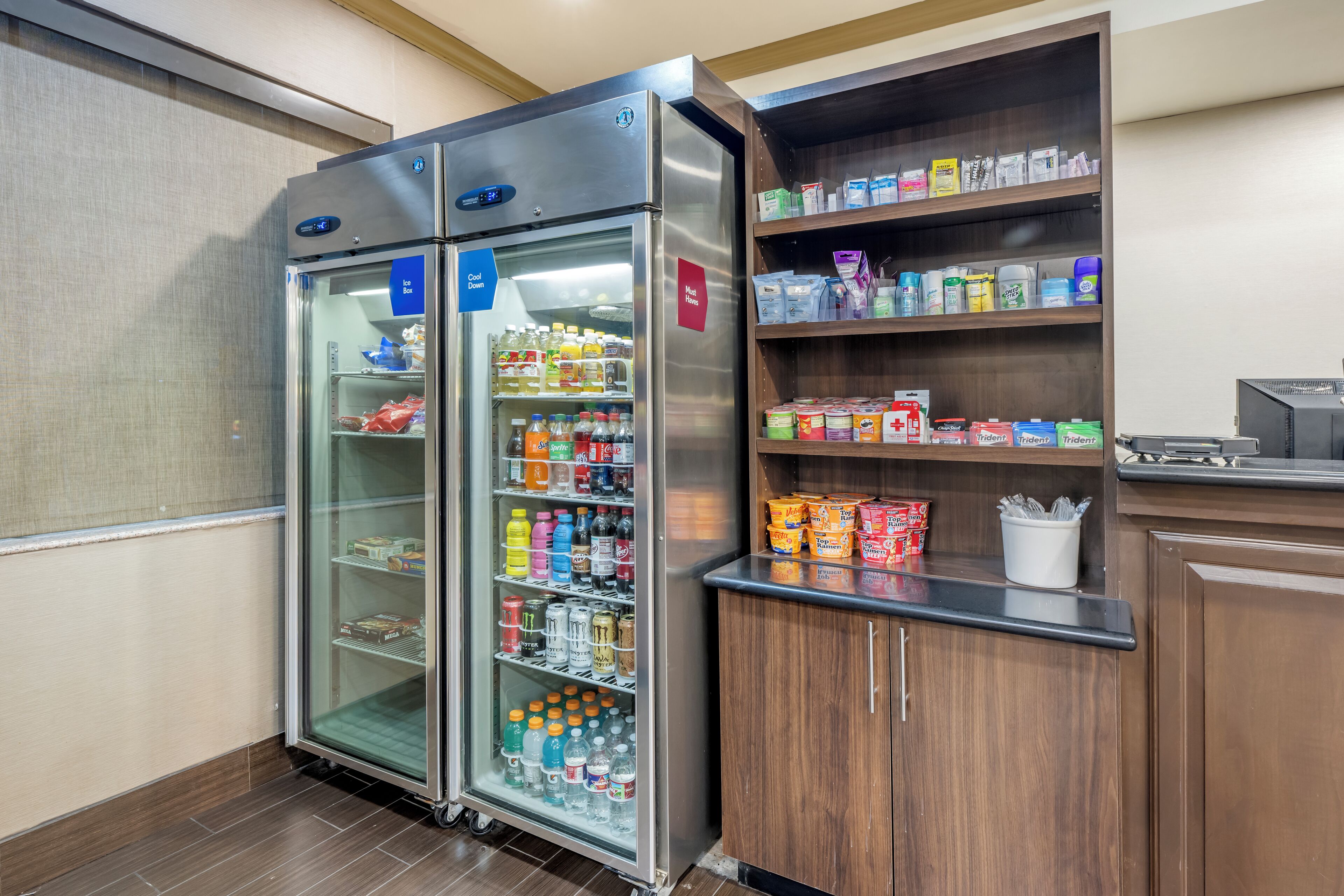 A commercial kitchen with stainless steel appliances, including two refrigerators and a wooden cabinet with shelves stocked with food items.