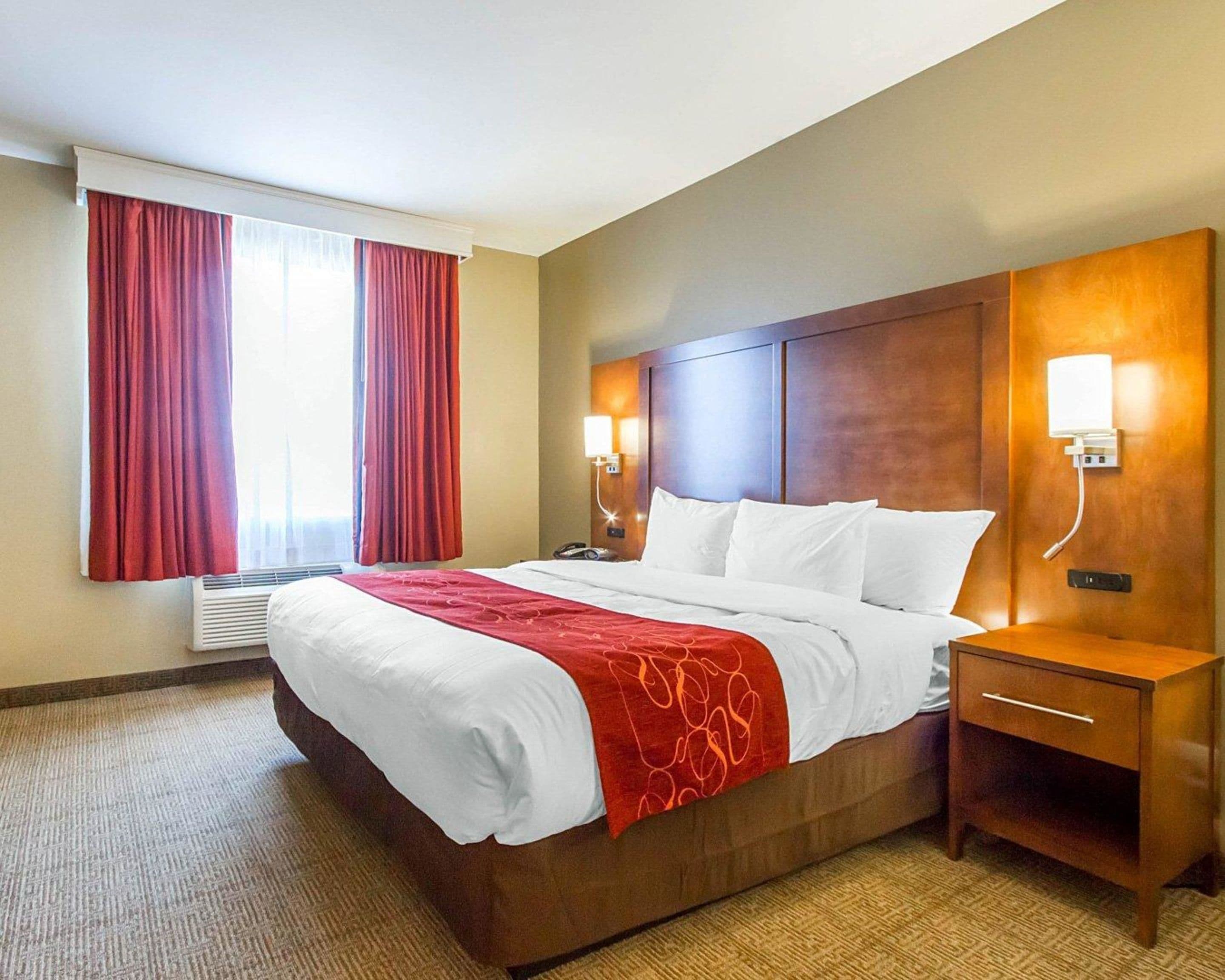 A hotel room with a large bed, red curtains, a wooden headboard, bedside lamps, and a small wooden nightstand.