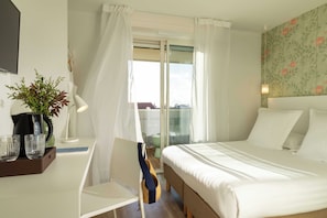 A hotel room with a bed, a desk with a lamp, a chair, a potted plant, and a view of the outside.