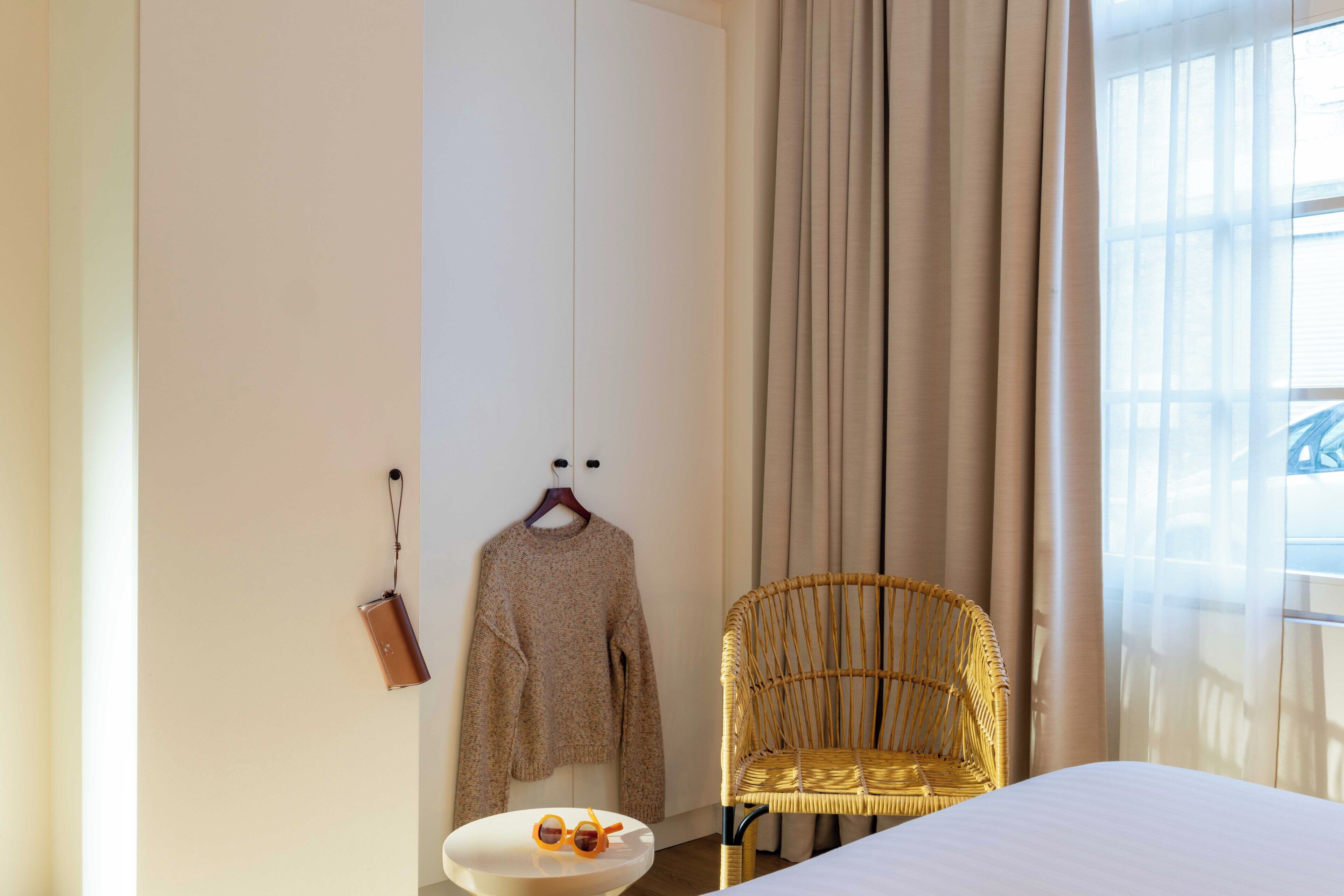 Un dormitorio con una cama, una silla de mimbre, un bolso colgado y un suéter en la pared.