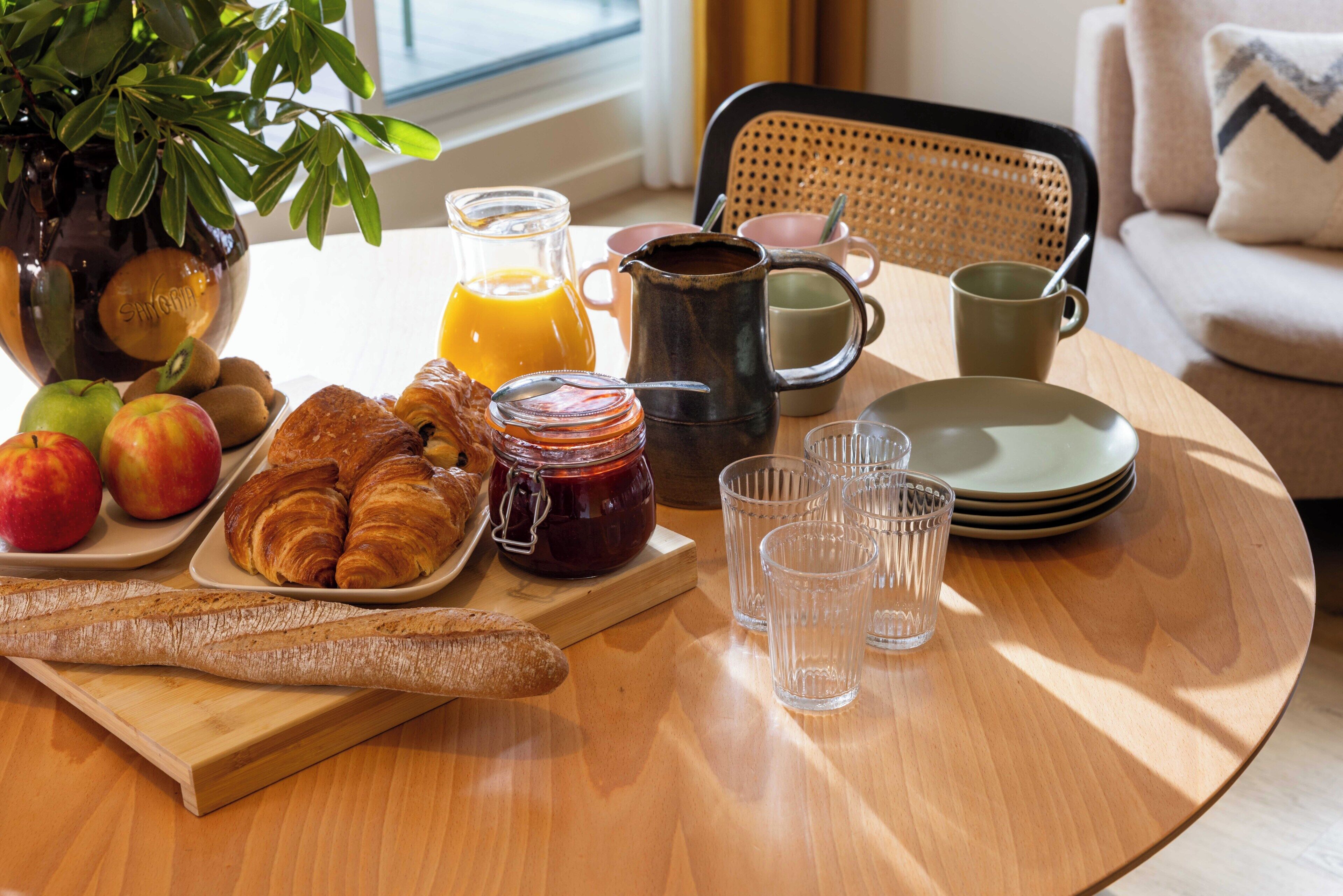 A breakfast setting with croissants, apples, a jar of jam, a pitcher of orange juice, and a coffee pot on a wooden table.