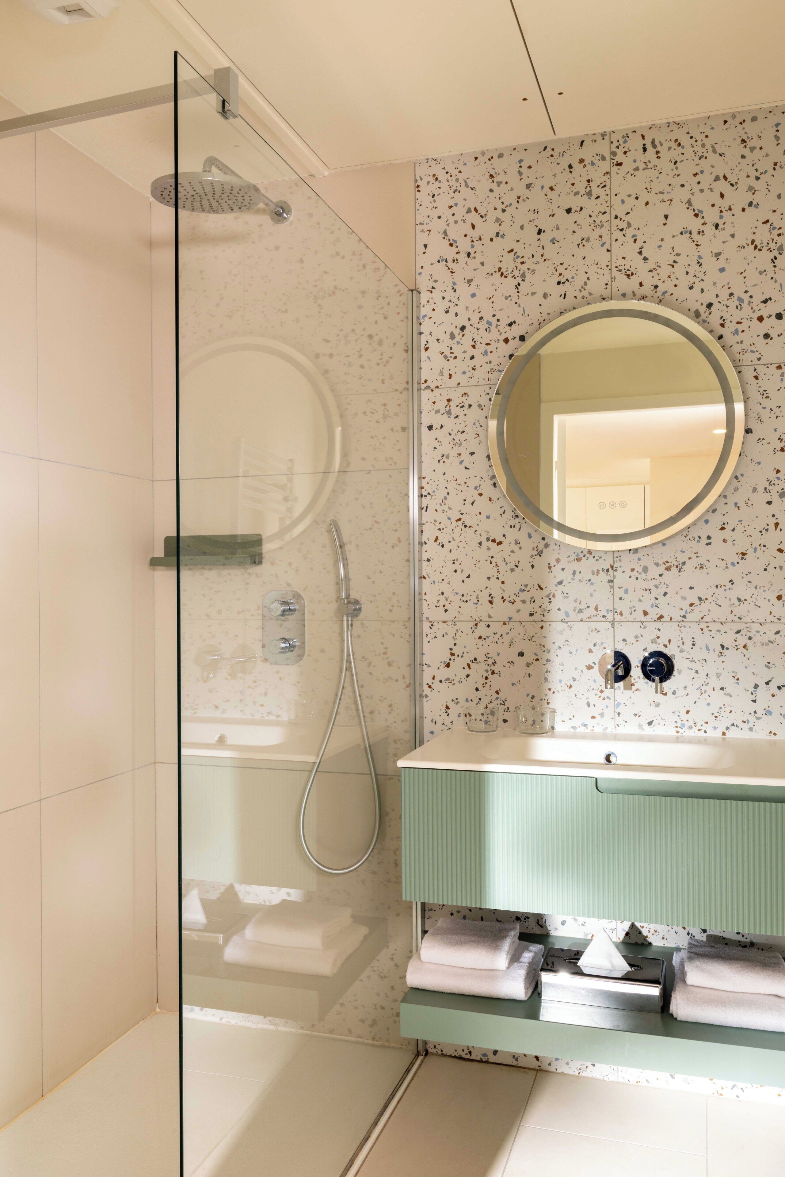 Un baño moderno con cabina de ducha de vidrio, un espejo redondo y un lavabo blanco con encimera verde.
