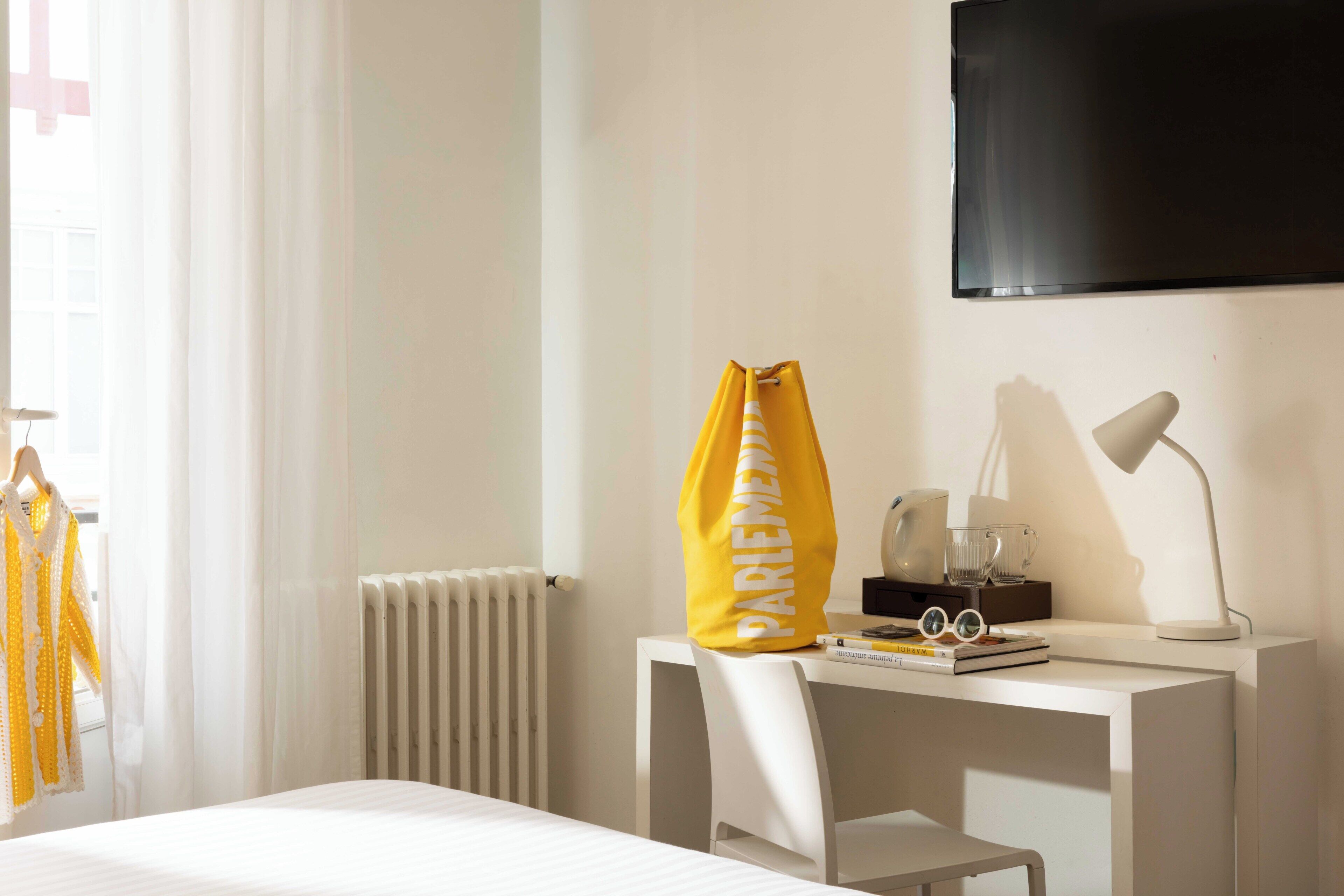 Una habitación de hotel con cama, escritorio con silla, televisión, radiador y ventana con cortinas.