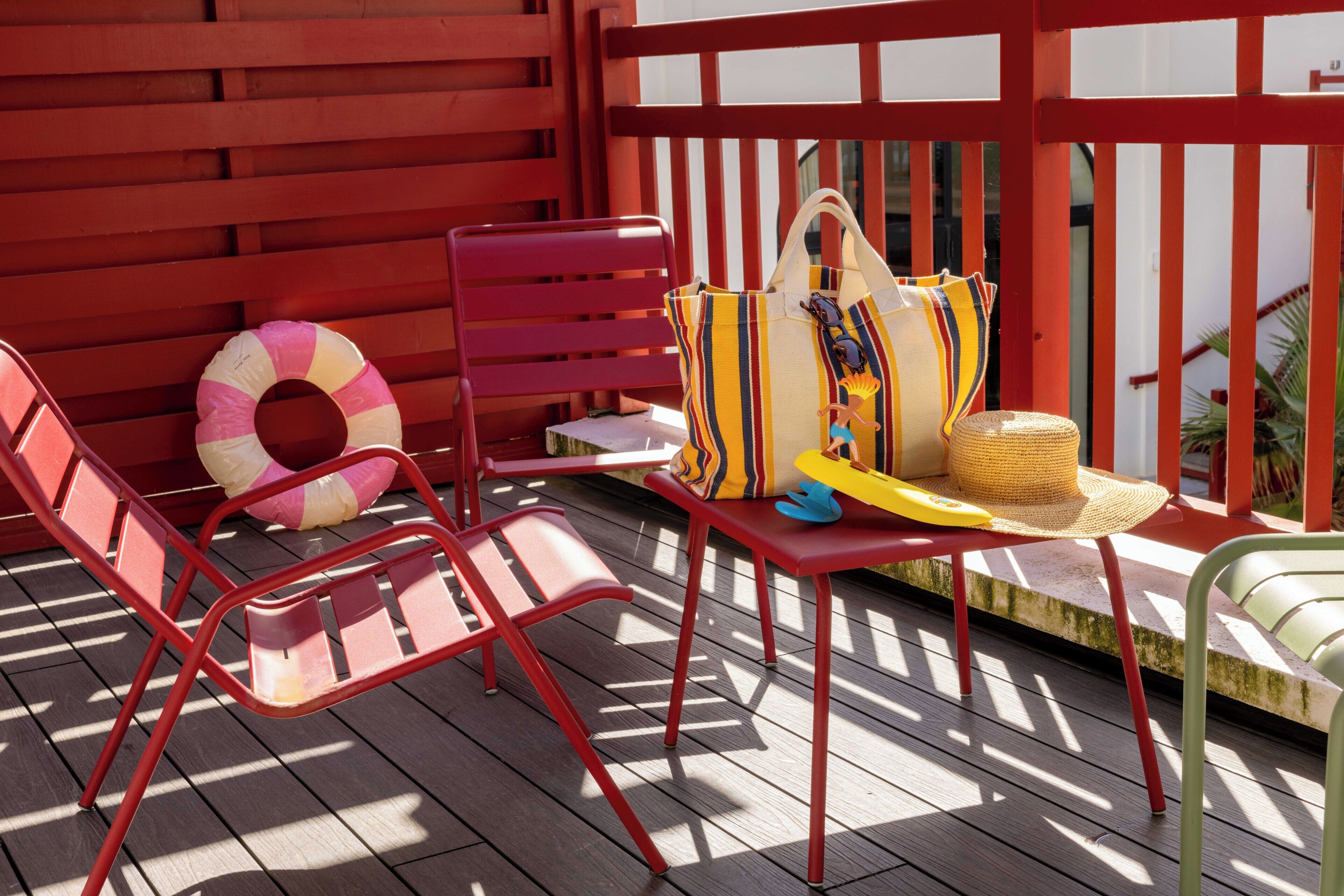 A red-framed outdoor seating area with a sun lounger, a beach ball, and a straw hat on a wooden deck.