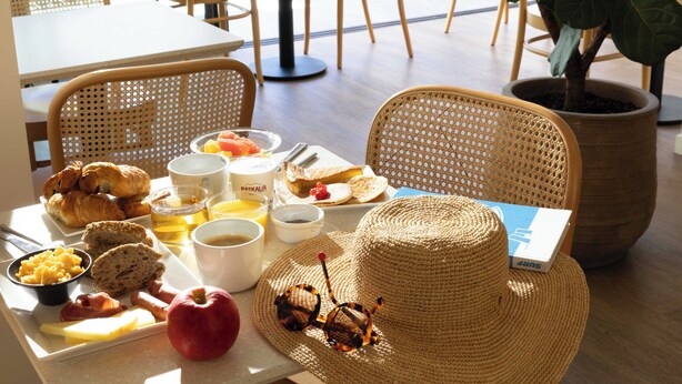 A breakfast tray with croissants, eggs, bacon, and fruit, a straw hat, and sunglasses on a table. - Hôtel Kemaris (Biarritz)