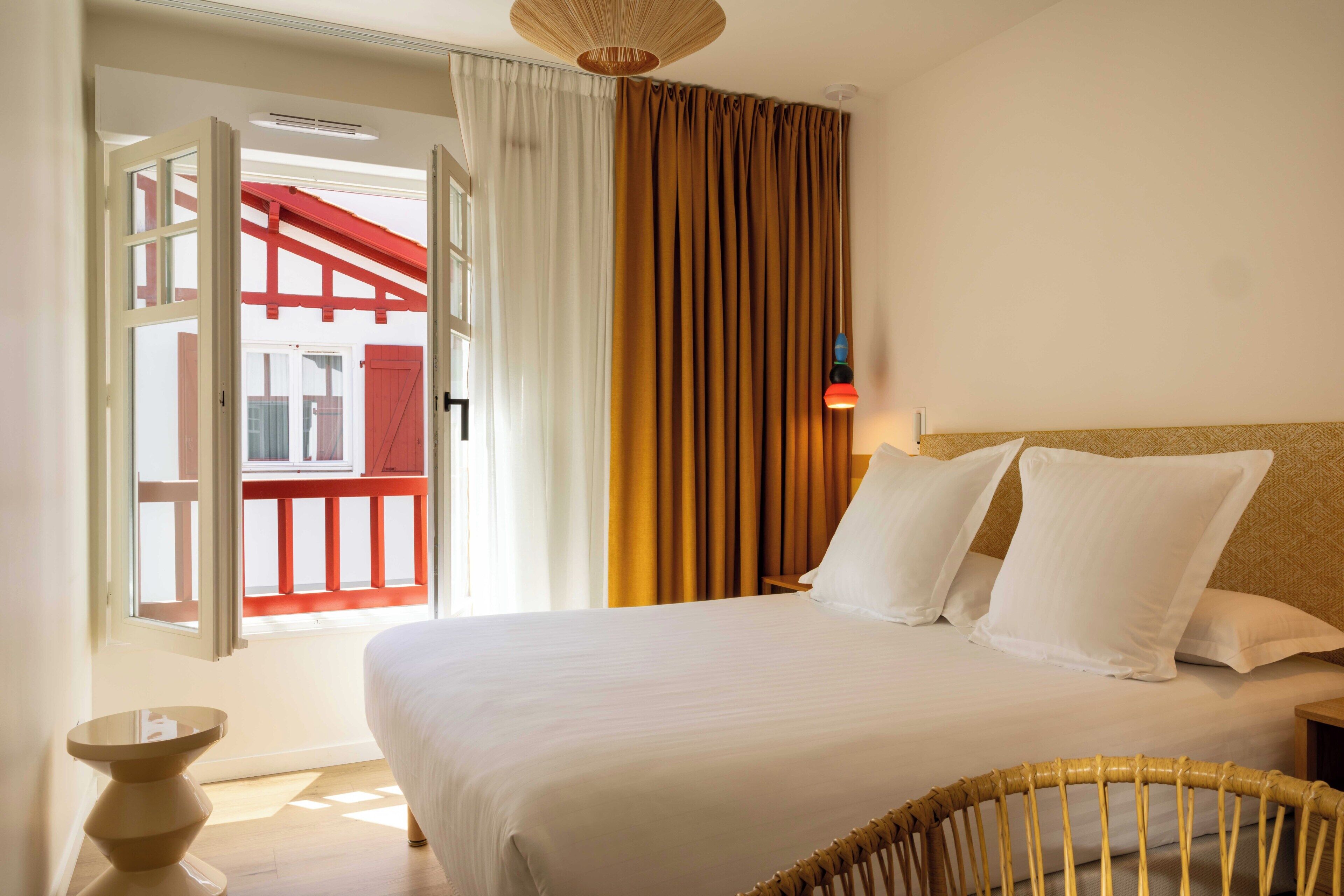 Een hotelkamer met een bed, nachtkastje, lamp en uitzicht op een rood balkon.