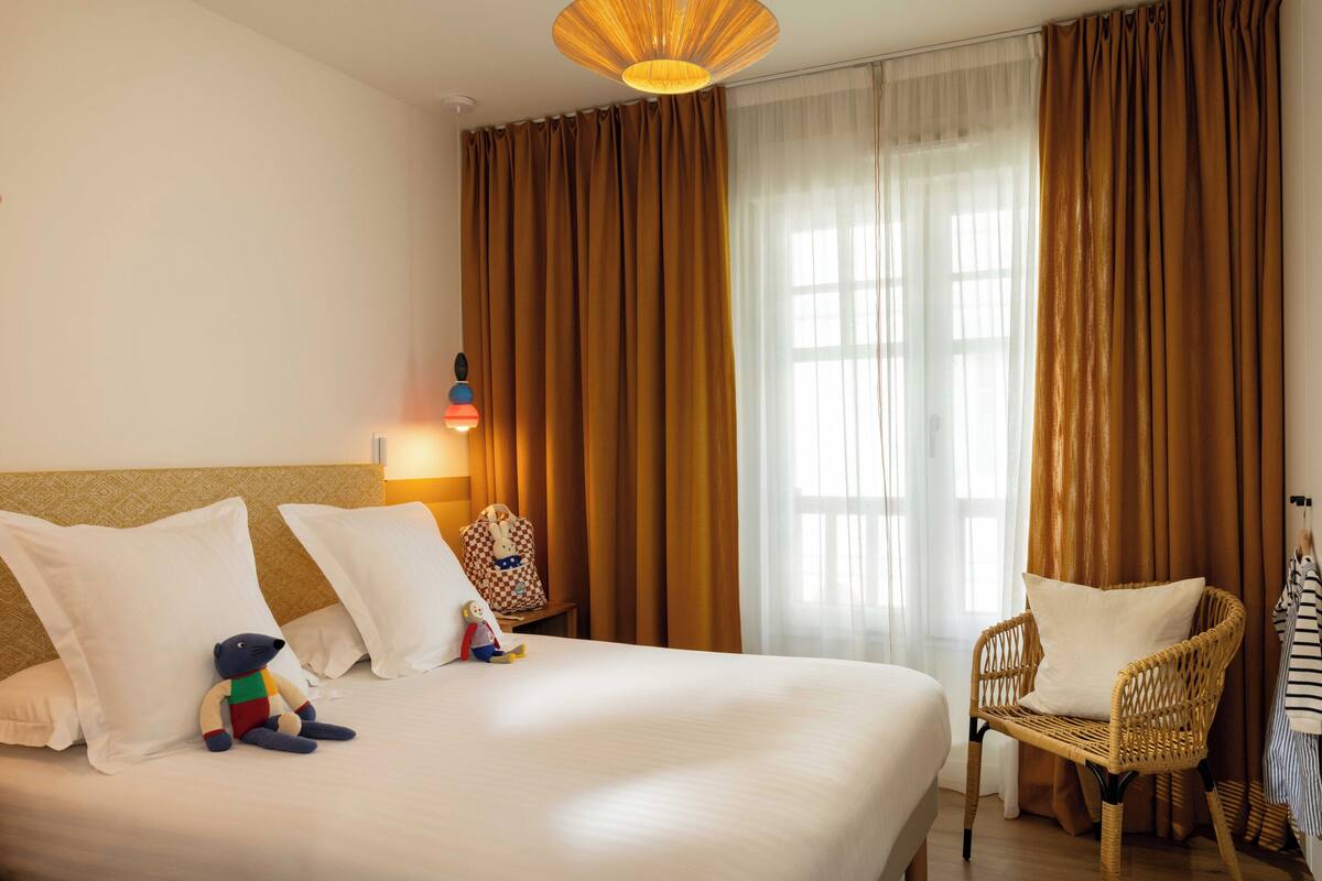 A hotel room with a large bed, a wicker chair, a small table with a lamp, and a view of the outside through sheer curtains.