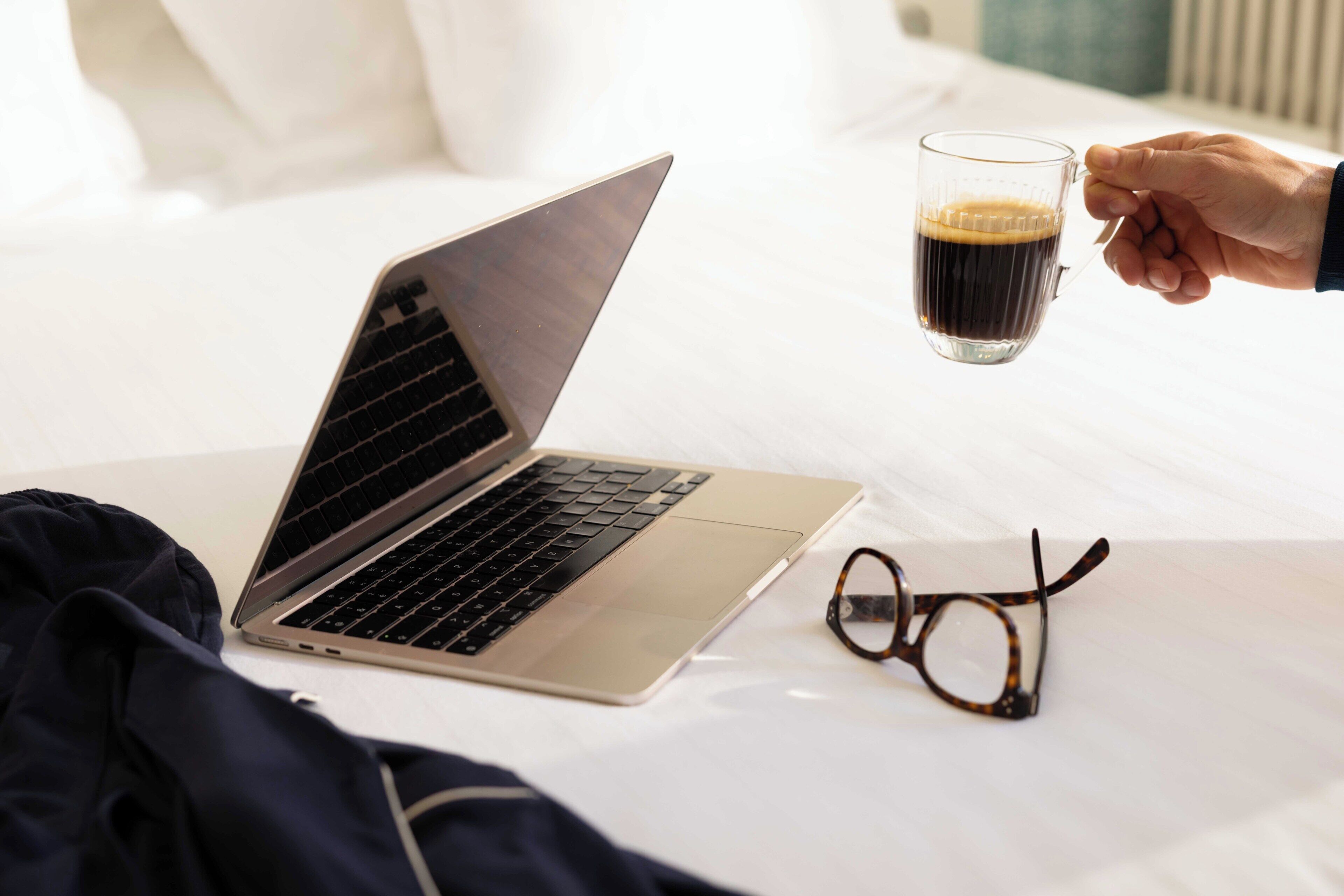 A laptop on a bed with a pair of glasses and a hand holding a mug.