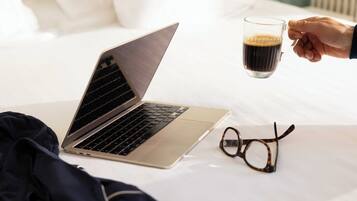 A laptop on a bed with a pair of glasses and a hand holding a mug.