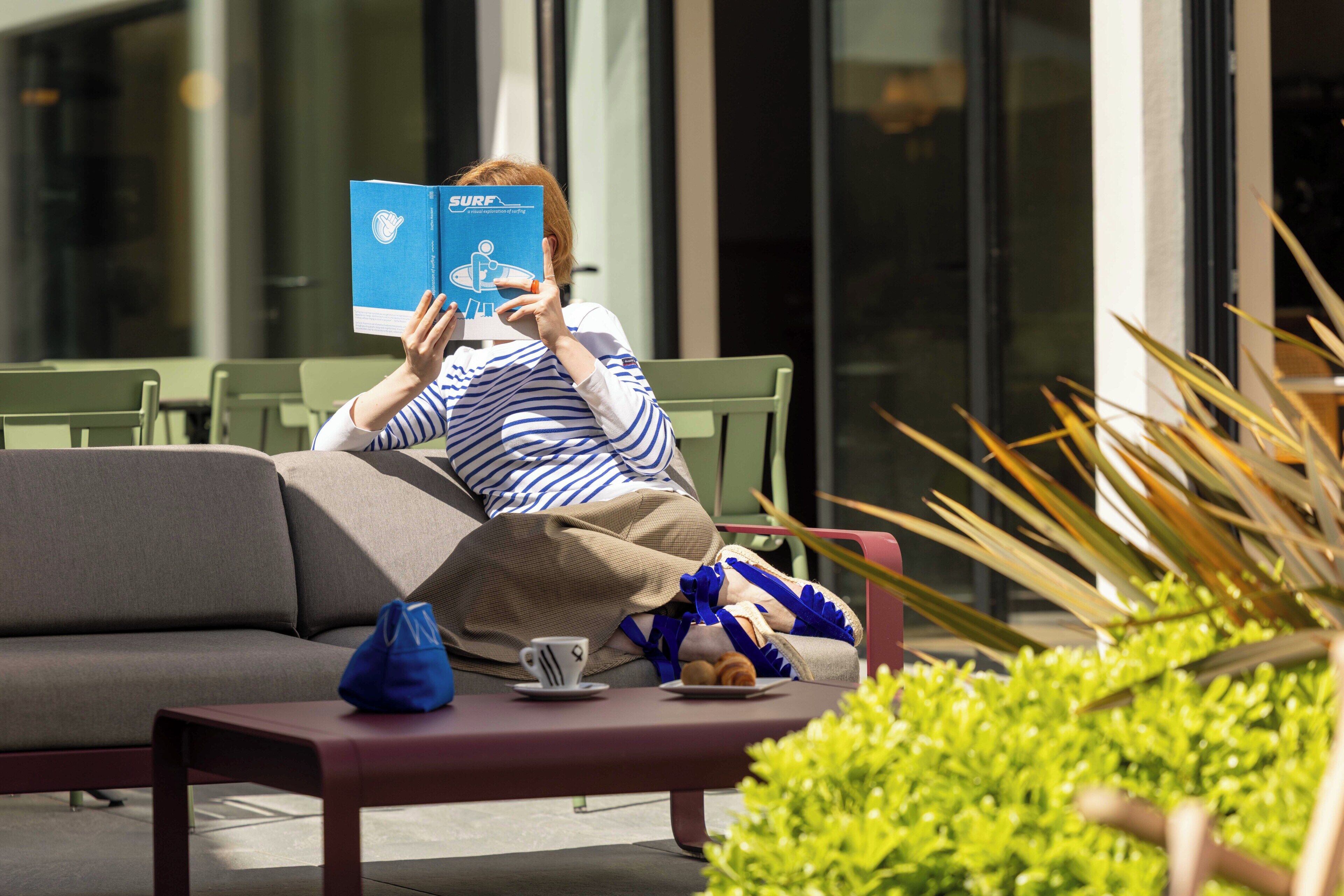 A person reading a book on a bench with a cup and pastries nearby, in an outdoor seating area with greenery and modern architecture in the background.