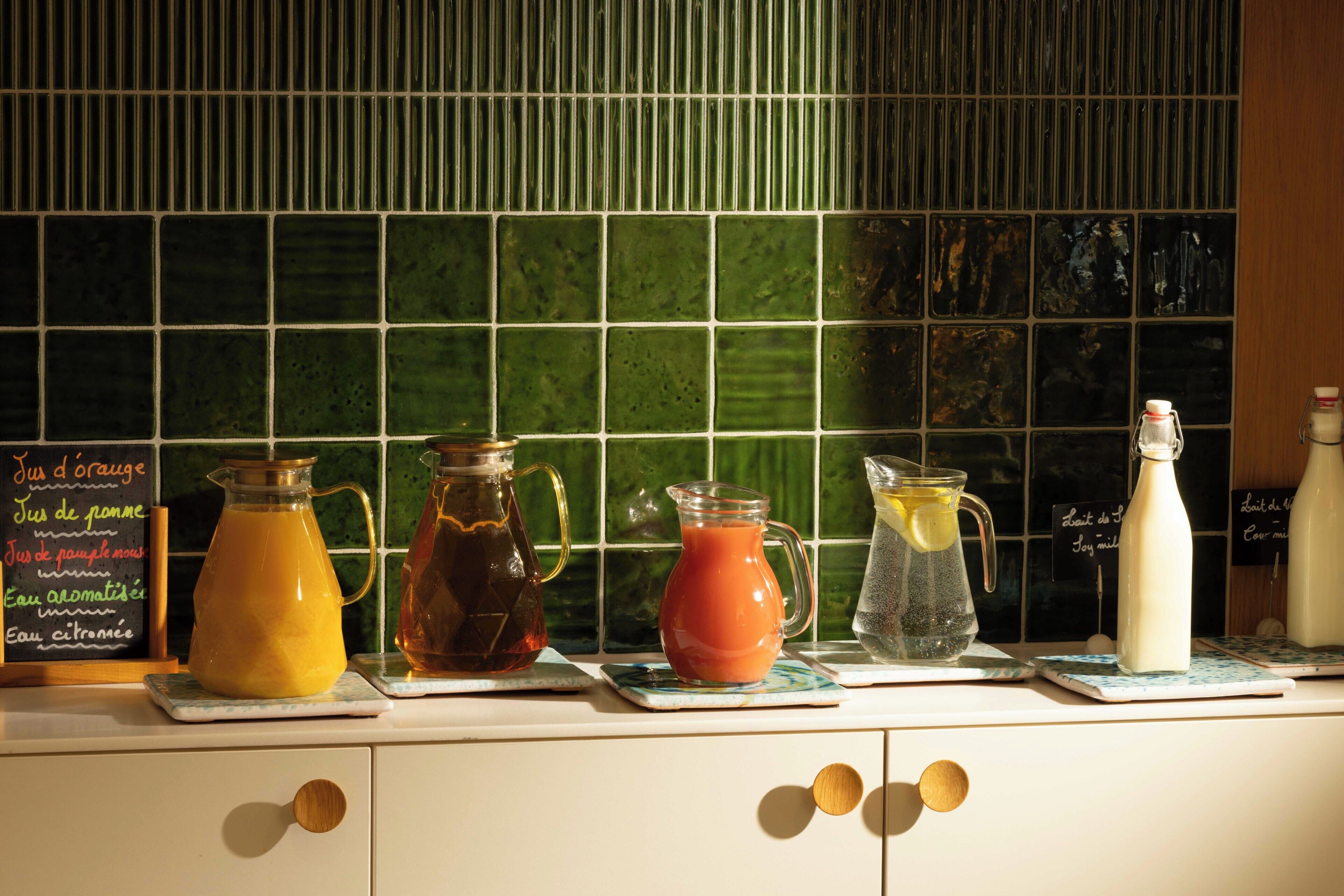 Un comptoir de cuisine sur lequel sont disposés divers récipients en verre contenant différentes boissons, un tableau noir servant de menu et un pare-feu vert en carreaux.