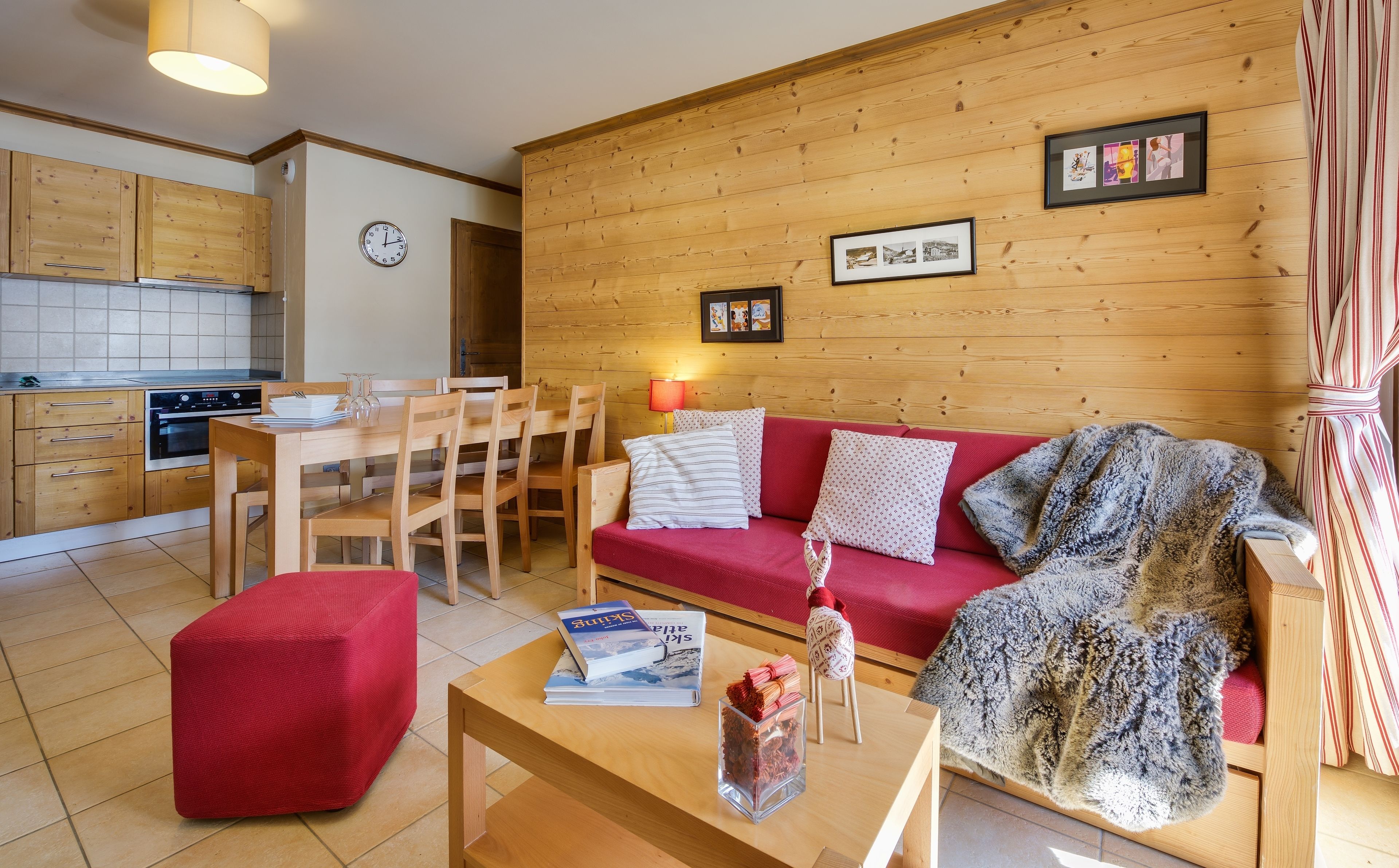 A cozy living room with a red sofa, wooden dining table, and a kitchen in the background.
