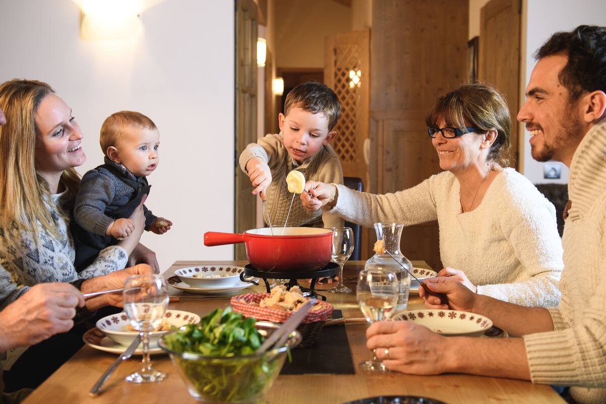Eine Familie isst gemeinsam an einem Holztisch. Auf dem Tisch stehen ein roter Topf, Teller und Gläser.