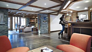 A spacious lobby with a high ceiling, stone walls, and wooden beams. There is a reception desk, seating area, and a view of the outside. - CGH Résidences & SPAS Le Ruitor (Sainte-Foy-Tarentaise)