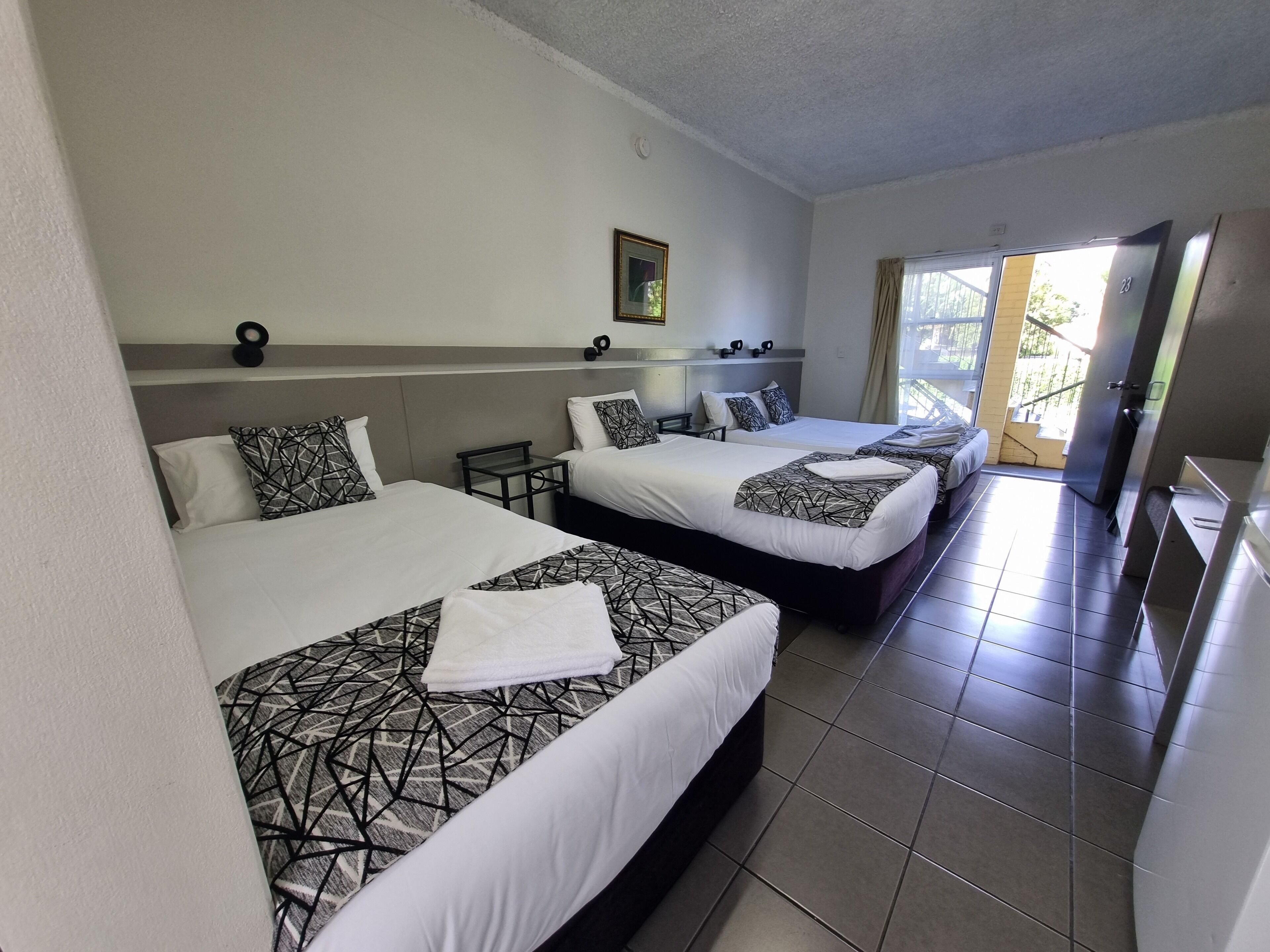 A hotel room with three beds, each with a geometric-patterned bedspread and white pillows. There is a small table between the beds, and a framed picture on the wall. The room has a tiled floor and a sliding glass door leading to an outdoor area.