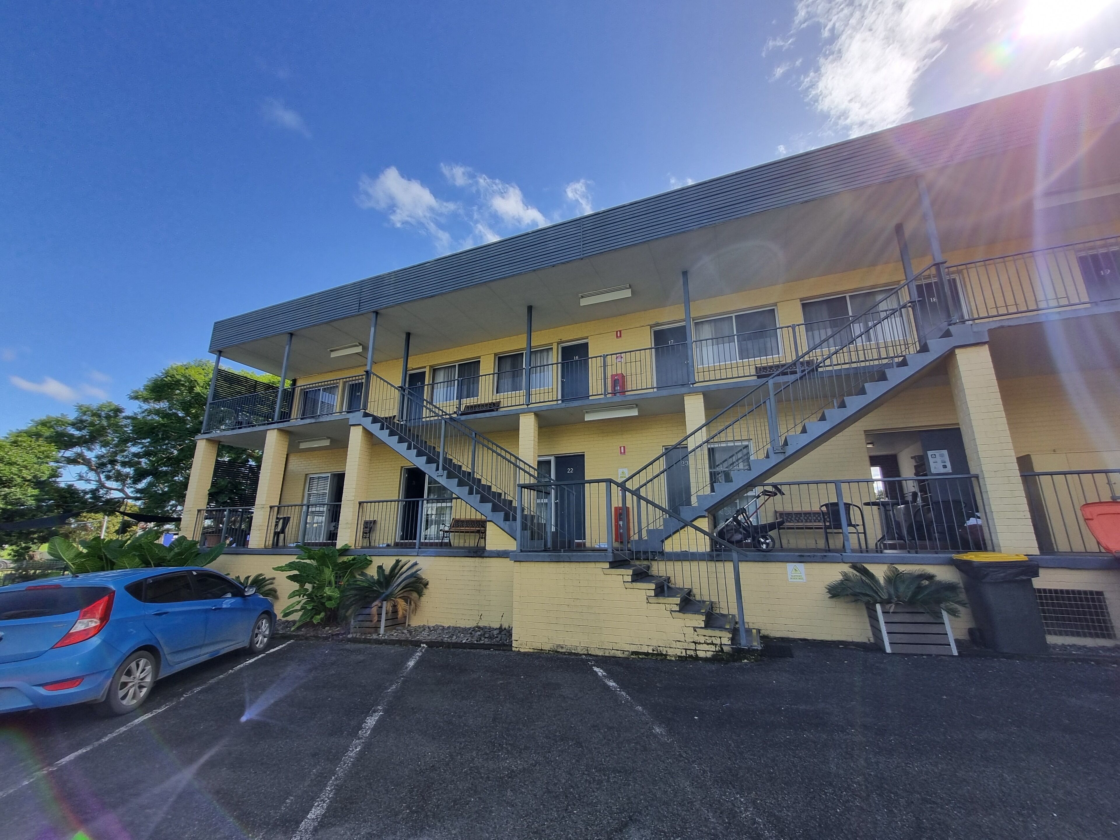 A multi-story building with a yellow facade, multiple balconies, and a parking area with a blue car.