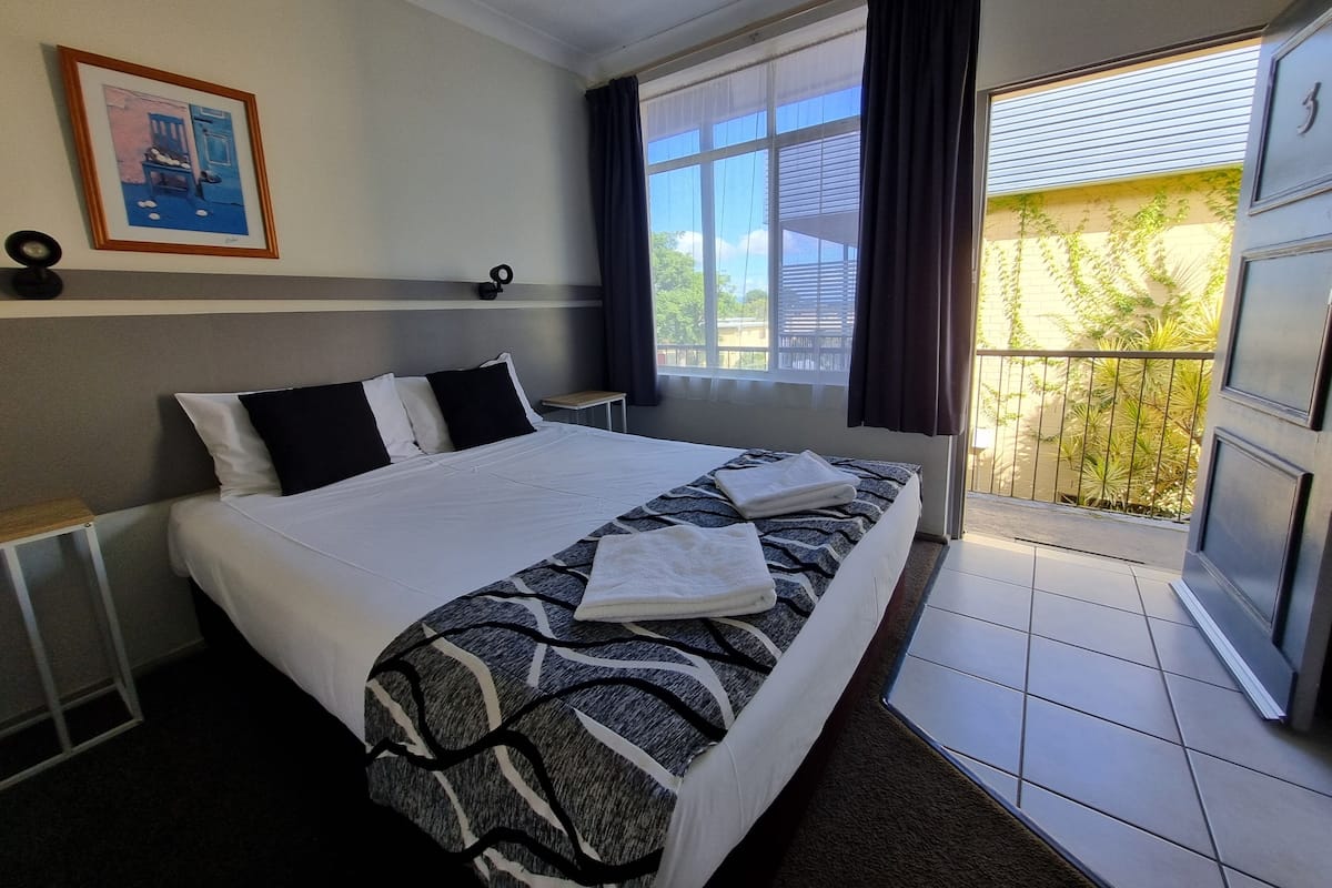A hotel room with a large bed, a small bedside table, a window with curtains, and a balcony with a view of greenery.