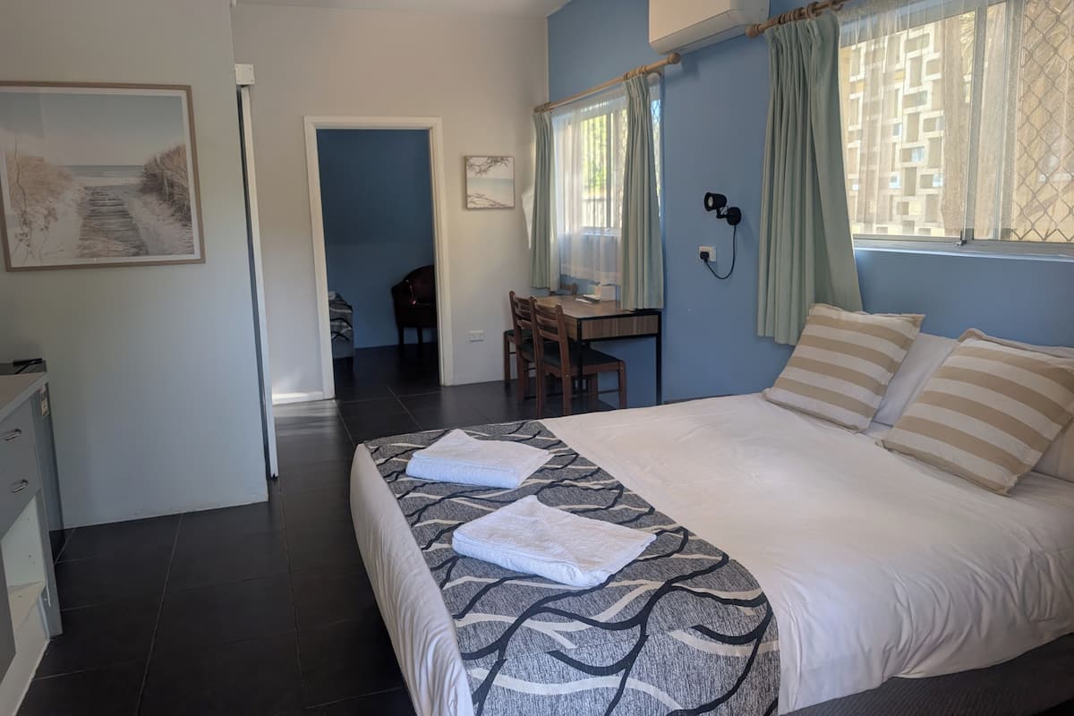 A bedroom with a bed, a desk, and a chair. There is a window with curtains and an air conditioning unit on the wall.