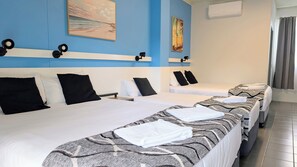 Habitación de hotel con tres camas, cada una con cubrecama blanco y almohadas negras. Hay lámparas de pared en el cabecero y un cuadro sobre las camas.