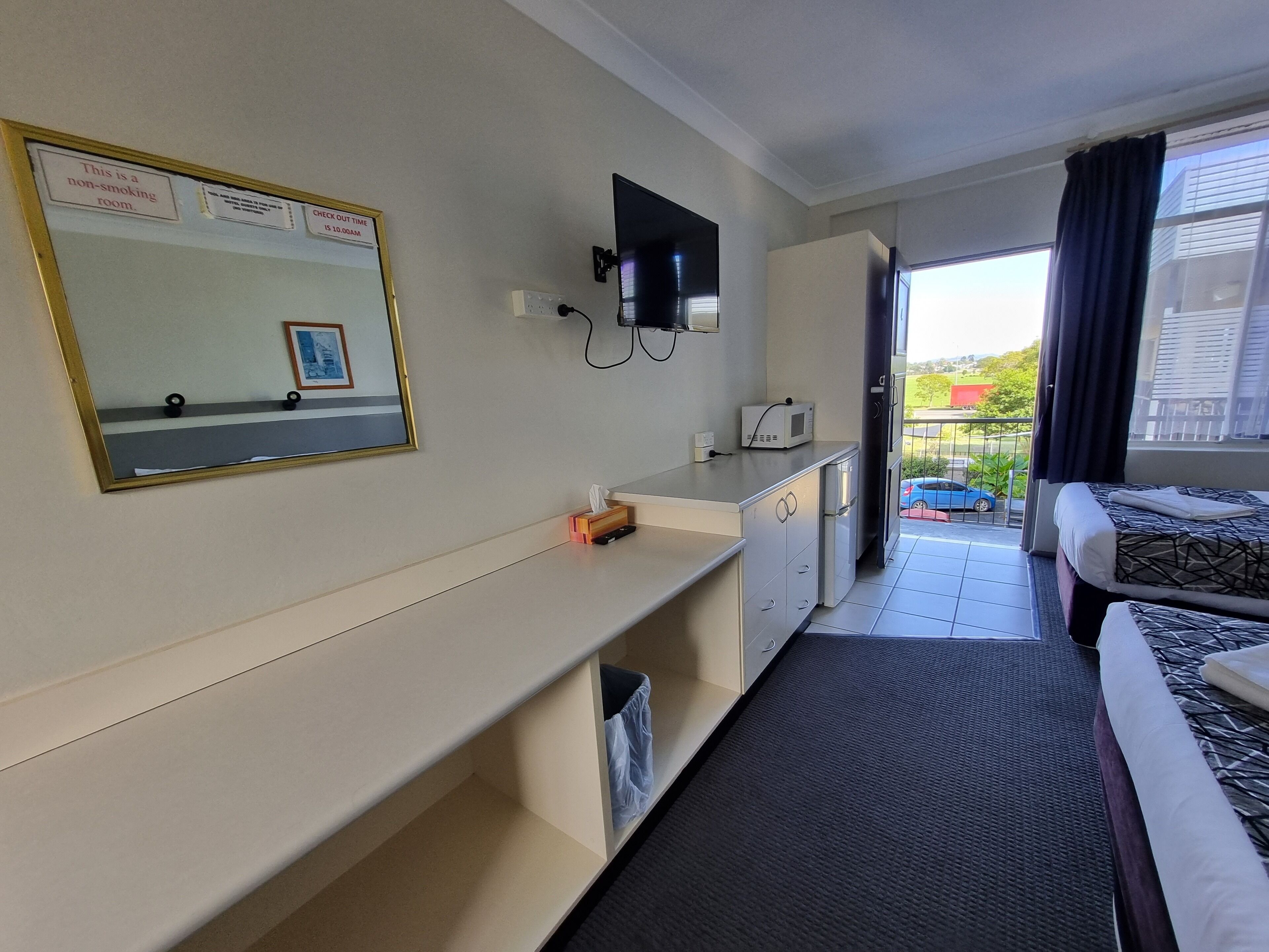 A hotel room with a check-out time sign, a non-smoking room notice, a flat-screen TV, a microwave, a small desk with a tissue box, a bed with patterned bedding, and a view of the outside balcony.