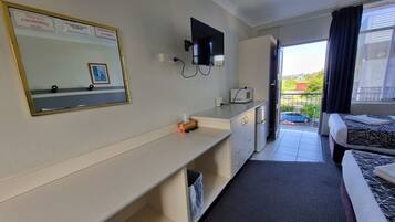 A hotel room with a check-out time sign, a non-smoking room notice, a flat-screen TV, a microwave, a small desk with a tissue box, a bed with patterned bedding, and a view of the outside balcony.