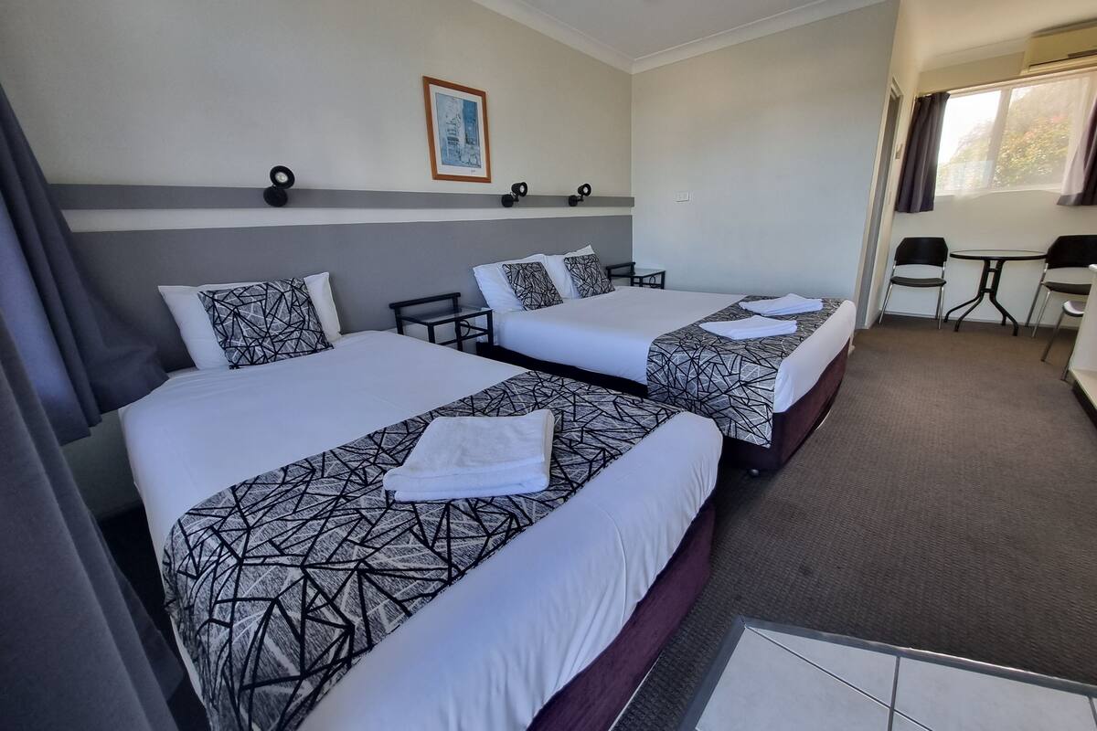 A hotel room with two beds, a small table, a chair, and a window.