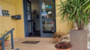 A building entrance with a yellow wall, a blue door, and a staircase leading inside. There are potted plants and a metal grate at the entrance.