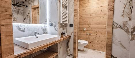 Un baño moderno con tocador de madera, baldosas de mármol, lavamanos blanco y puerta de madera.