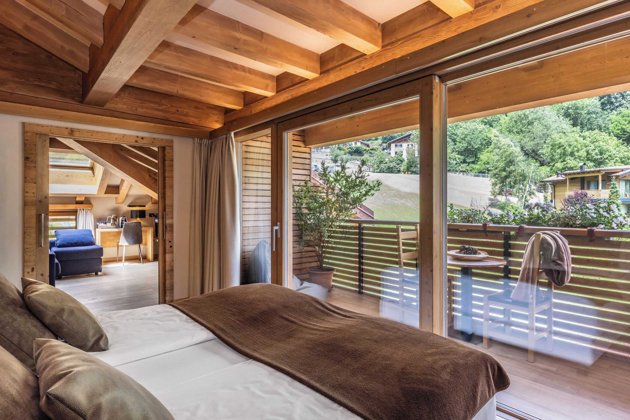 Quarto com uma cama grande, vigas de madeira no teto e uma porta de vidro deslizante para uma varanda com vista para casas e vegetação.