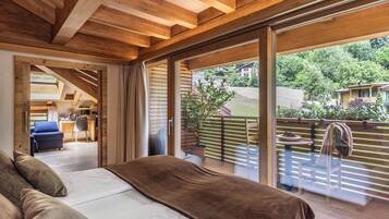 Quarto com uma cama grande, vigas de madeira no teto e uma porta de vidro deslizante para uma varanda com vista para casas e vegetação.