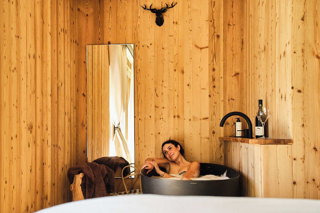 A wooden cabin interior with a bathtub, a wooden shelf with bottles, and a deer head mounted on the wall.