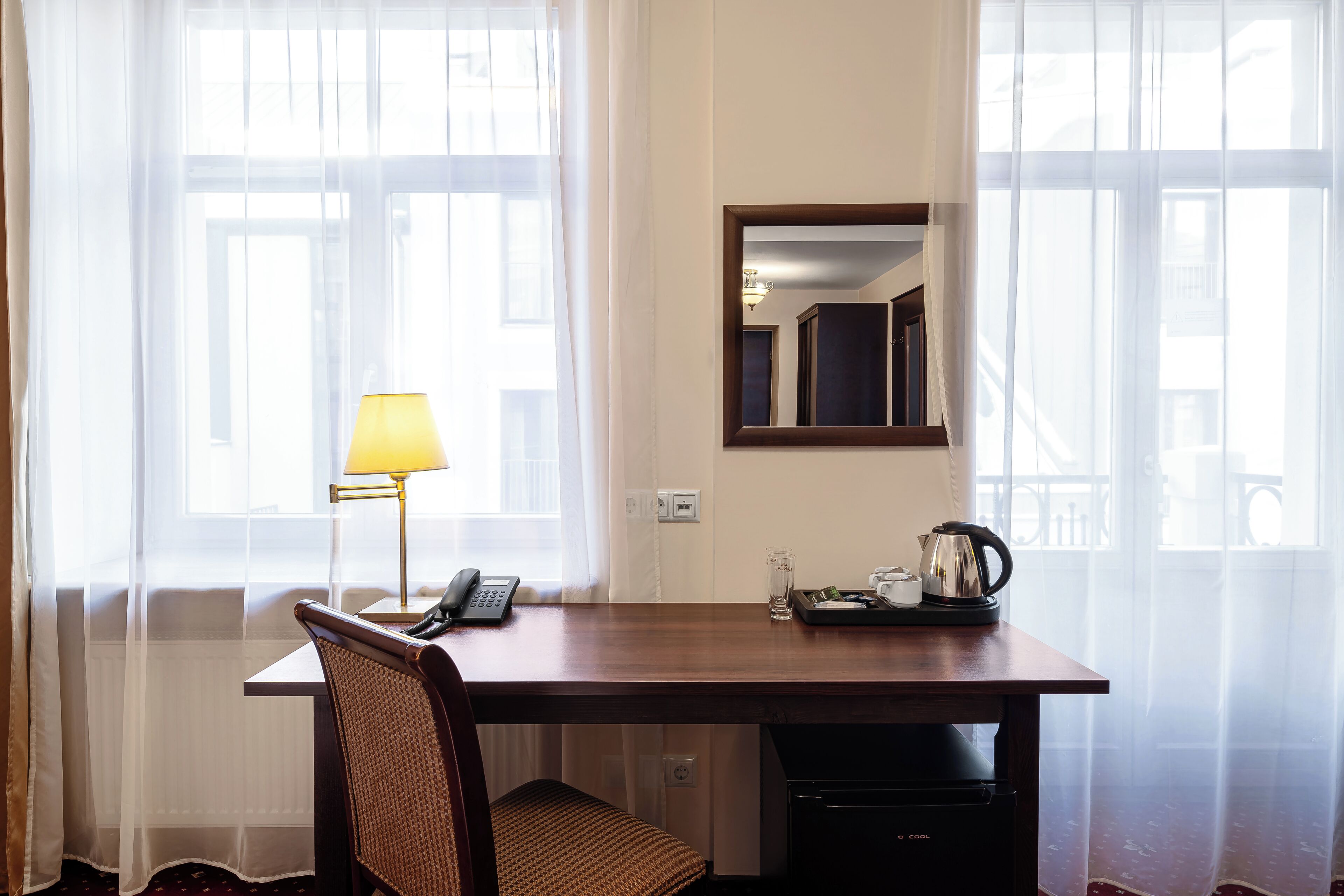 Habitación de hotel con un escritorio de madera, una silla, una lámpara, un teléfono, una cafetera y un vaso de agua.