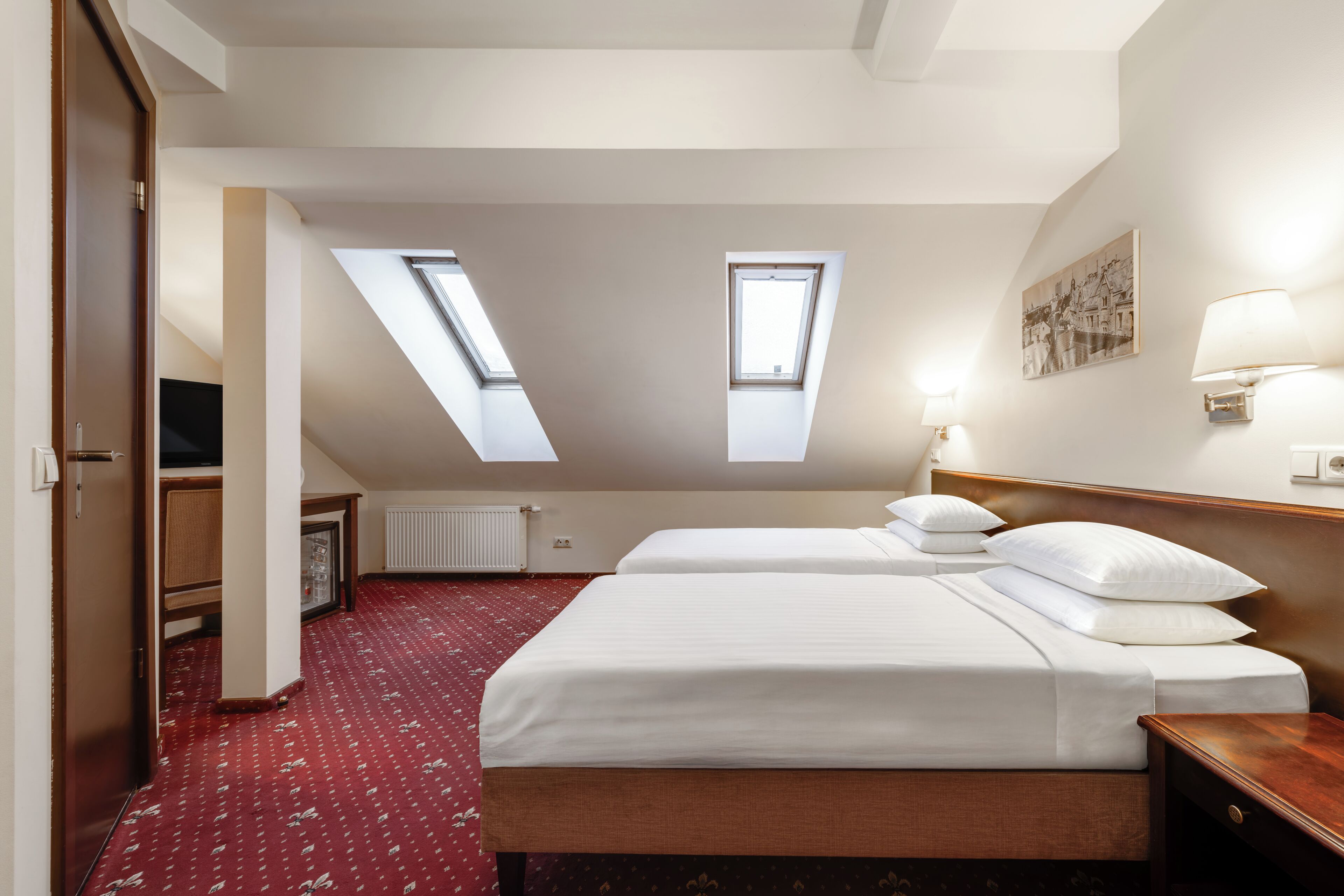 A hotel room with two beds, skylights, a wooden headboard, a bedside lamp, a radiator, and a wall-mounted picture.