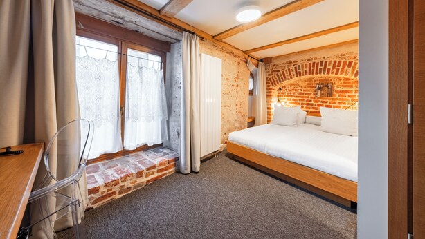 Una habitación con pared de ladrillo, cama de madera, ventana con cortinas y un escritorio con silla.