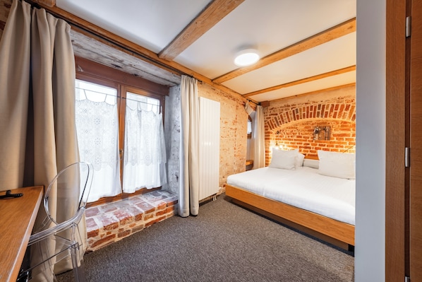 Una habitación con pared de ladrillo, cama de madera, ventana con cortinas y un escritorio con silla.