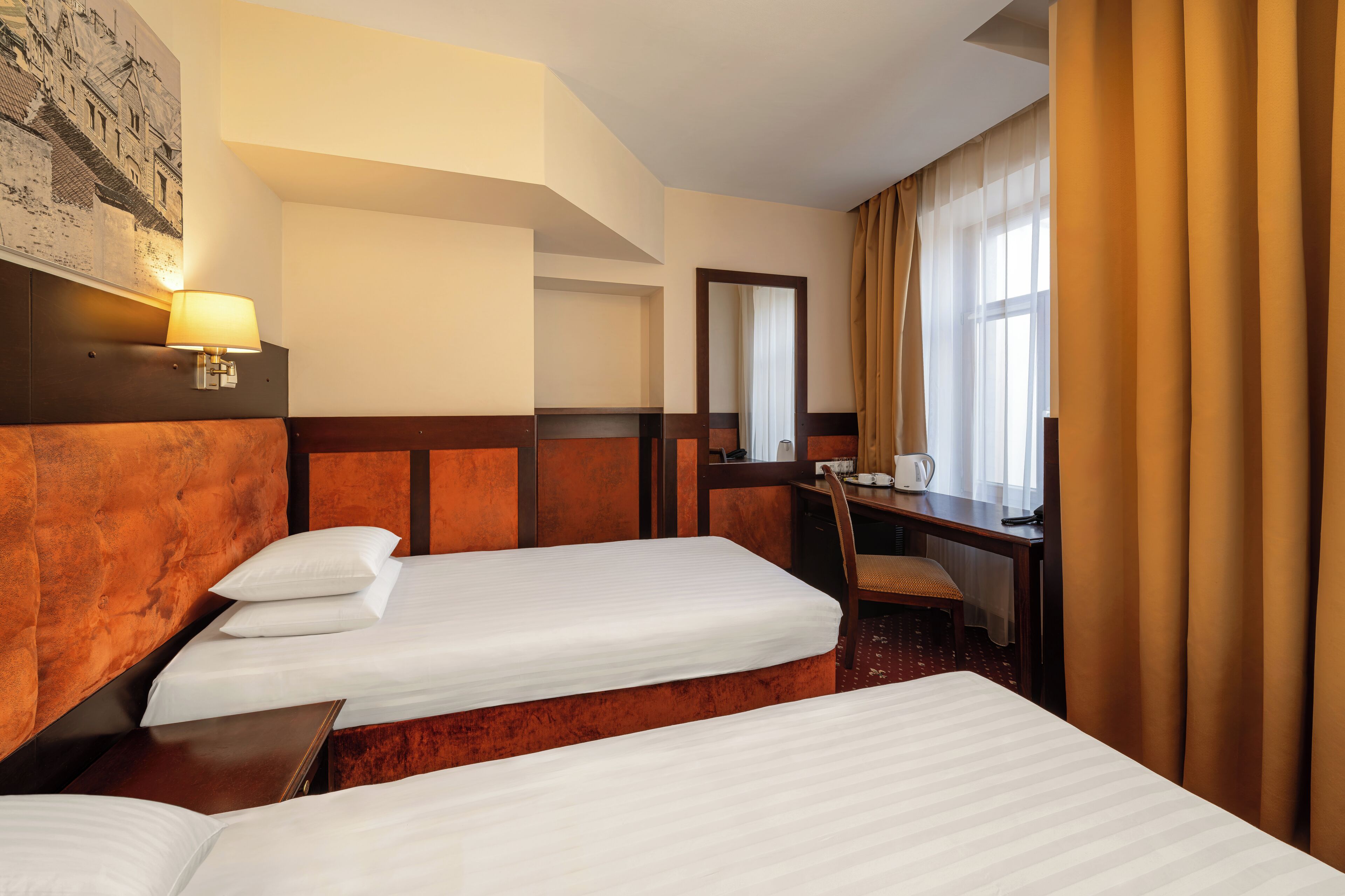 Habitación de hotel con dos camas, un escritorio, una silla, una lámpara y un ventanal grande con cortinas.
