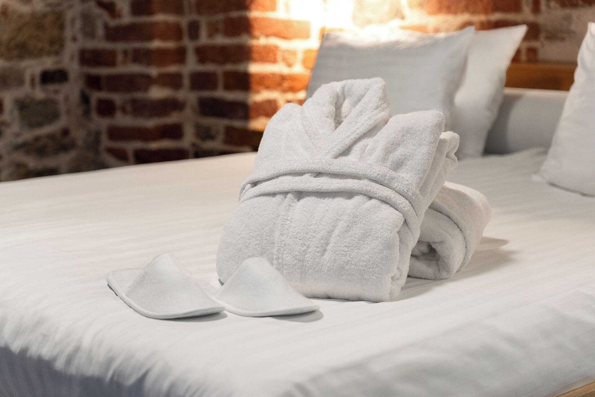 A neatly made bed with a folded towel and a pair of slippers on a white bedspread, set against a brick wall.
